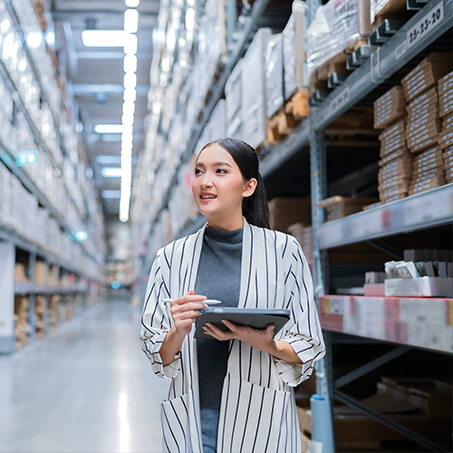 Smiling woman walking in warehouse aisle with mobile device.